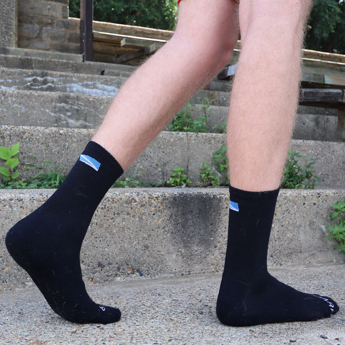 A person wearing black crew socks with small blue and white logo tabs near the top, standing on concrete outdoor steps.